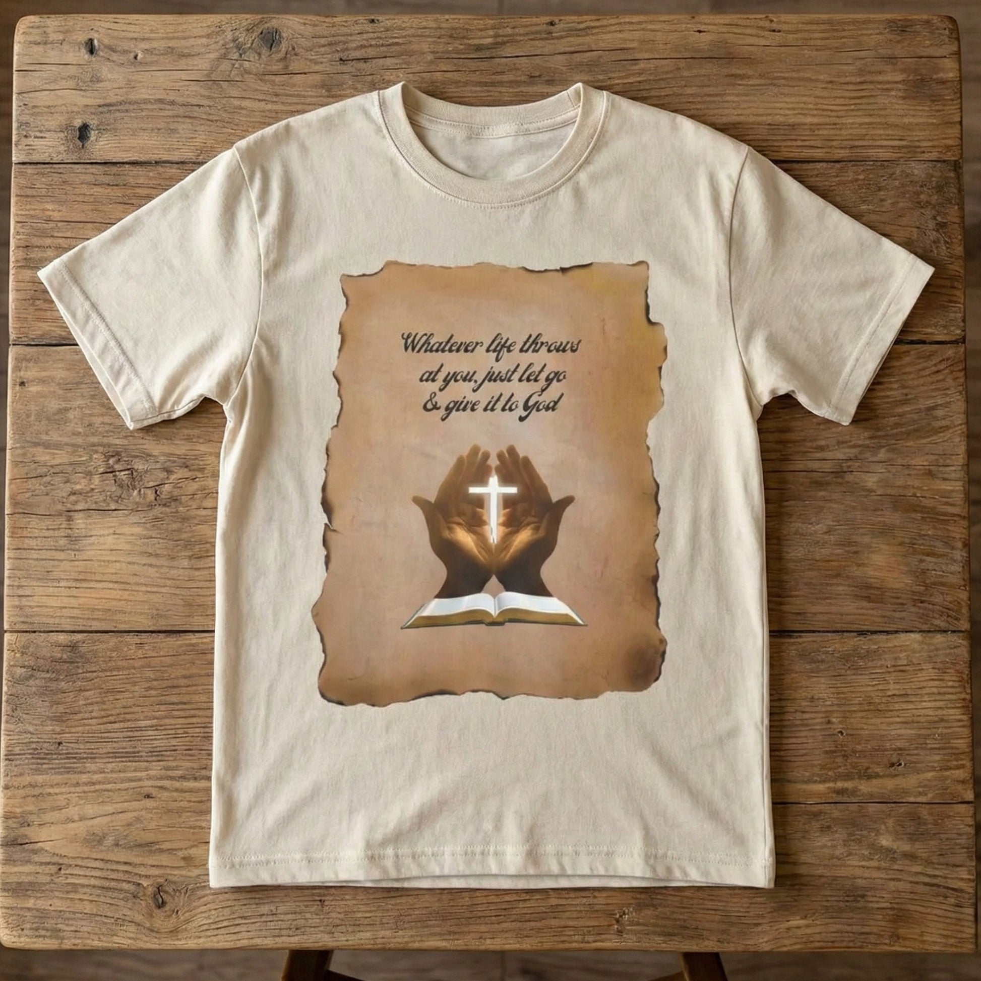 Cream t-shirt with "Give It To God" parchment graphic laid flat on a rustic wooden table as a spiritual encouragement gift by Everyday Rebel Co.